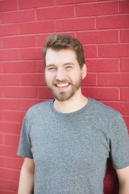 Zach smiling and standing up against a bright red brick wall, wearing a static patterned gray shirt